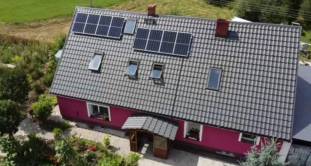 Widok z góry na dom z szarą dachówką i panelami słonecznymi, otoczony zielenią, z widocznym ogrodem i fragmentem pola.