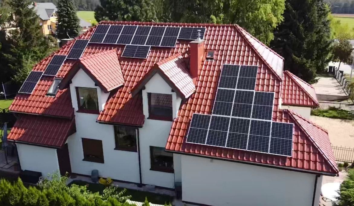 Widok z góry na dom jednorodzinny z czerwonym dachem, na którym zamontowane są panele słoneczne w różnych konfiguracjach, otoczony zielenią drzew.