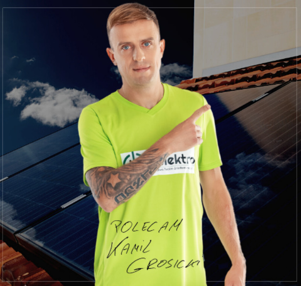 Mężczyzna w zielonej koszulce z logo firmy na tle paneli słonecznych i nieba, polecający usługi.