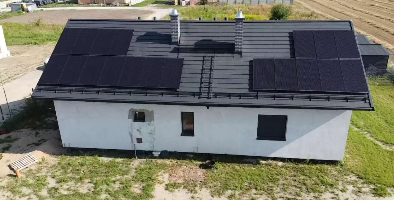 Dom jednorodzinny z zainstalowanymi panelami słonecznymi na dachu z blachodachówki, widoczne dwa kominy i drabina dachowa, elewacja budynku w trakcie wykończenia.