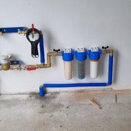 BUD-WOL Usługi Remontowo-Budowlane Daniel Wolski - Zamontowany system filtrów wody z manometrem i zaworami, rury miedziane i plastikowe, na białej ścianie w garażu. Widoczne filtry mechaniczne i węglowe.