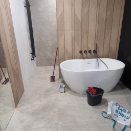 BUD-WOL Usługi Remontowo-Budowlane Daniel Wolski - Nowoczesna łazienka z wolnostojącą wanną, wykończona płytkami imitującymi drewno na ścianie. Widoczne akcesoria i narzędzia glazurnicze.