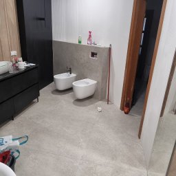 BUD-WOL Usługi Remontowo-Budowlane Daniel Wolski - Nowoczesna łazienka w trakcie wykańczania: biała armatura (bidet i toaleta), szare płytki podłogowe, czarne szafki i drewniane akcenty. Widoczne narzędzia i chemia.