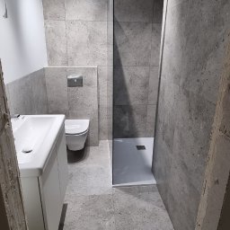 BUD-WOL Usługi Remontowo-Budowlane Daniel Wolski - Wykończona łazienka w szarościach z kabiną prysznicową typu walk-in, umywalką z szafką i podwieszaną toaletą. Widoczne płytki imitujące kamień i nowoczesne oświetlenie.