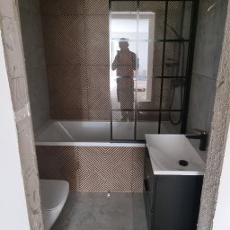 BUD-WOL Usługi Remontowo-Budowlane Daniel Wolski - Wykończona łazienka w Lęborku: geometryczne płytki na ścianie, czarna kabina prysznicowa, umywalka z szafką. Widok zza progu, w tle odbicie osoby.