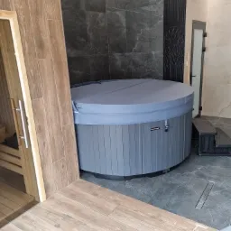 Nowoczesna łazienka z drewnianą sauną i szarą wanną jacuzzi z pokrywą, wykończona płytkami imitującymi drewno i marmur.