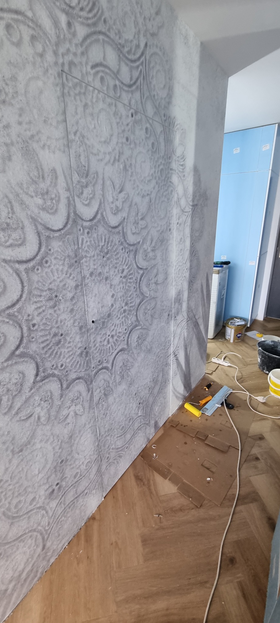 Ściana z dekoracyjną szarą strukturą imitującą wzór mandali, widoczny fragment podłogi z parkietu oraz narzędzia malarskie i zabezpieczenia podłogi w trakcie prac.