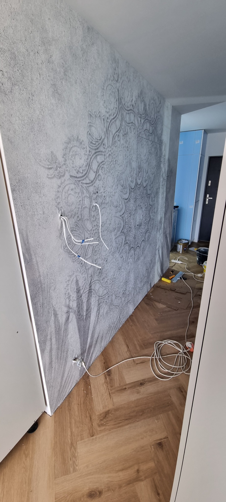 Ściana z dekoracyjnym wzorem w odcieniach szarości, widoczne kable elektryczne wystające ze ściany, podłoga wyłożona panelami w jodełkę, narzędzia i materiały budowlane w tle.