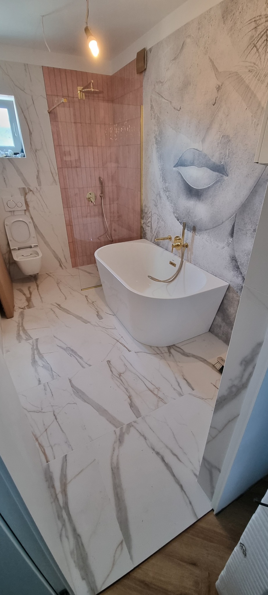 Nowoczesna łazienka z wolnostojącą wanną, złotą armaturą i marmurowymi płytkami, akcentowana ścianą prysznicową z różowych płytek oraz oryginalną tapetą z motywem twarzy.
