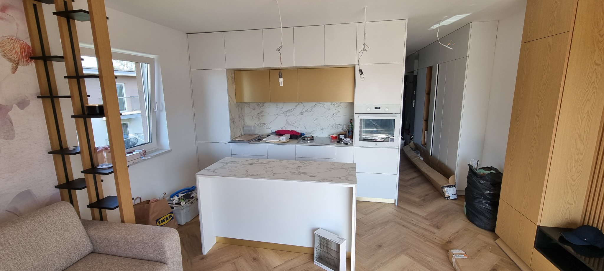 Nowoczesna kuchnia w trakcie wykańczania w Lęborku: białe szafki z elementami w kolorze złota, marmurowy blat, wyspa kuchenna i drewniana podłoga ułożona w jodełkę, widoczne kable od oświetlenia.