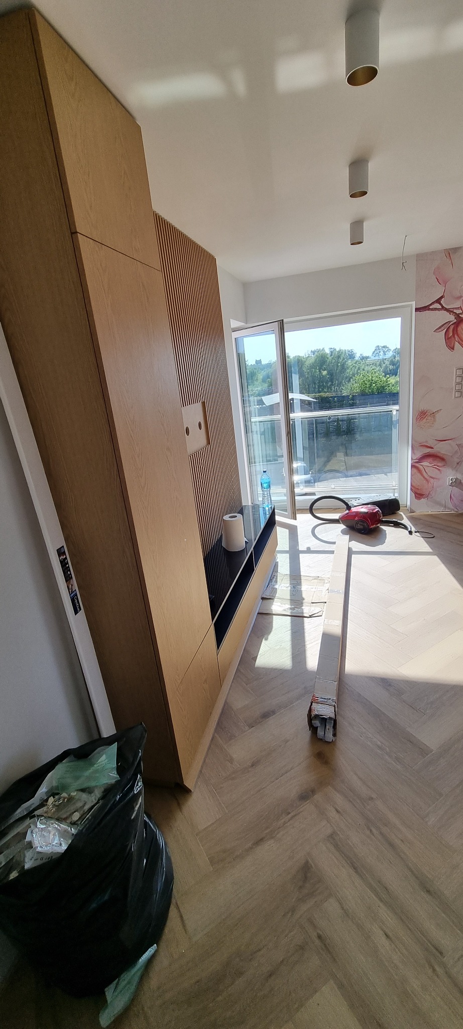 Montaż drewnianej zabudowy ściennej z lamelami i szafek w salonie, widoczny odkurzacz, karton i worek na śmieci na parkiecie ułożonym w jodełkę.
