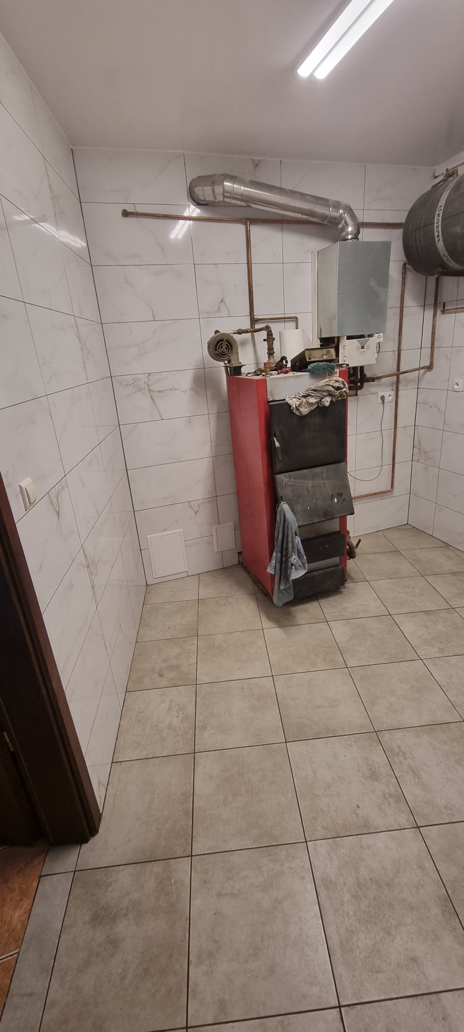 Kotłownia z piecem na paliwo stałe, widoczne rury miedziane, białe płytki na ścianach i podłodze, szara szmatka zwisająca z pieca.