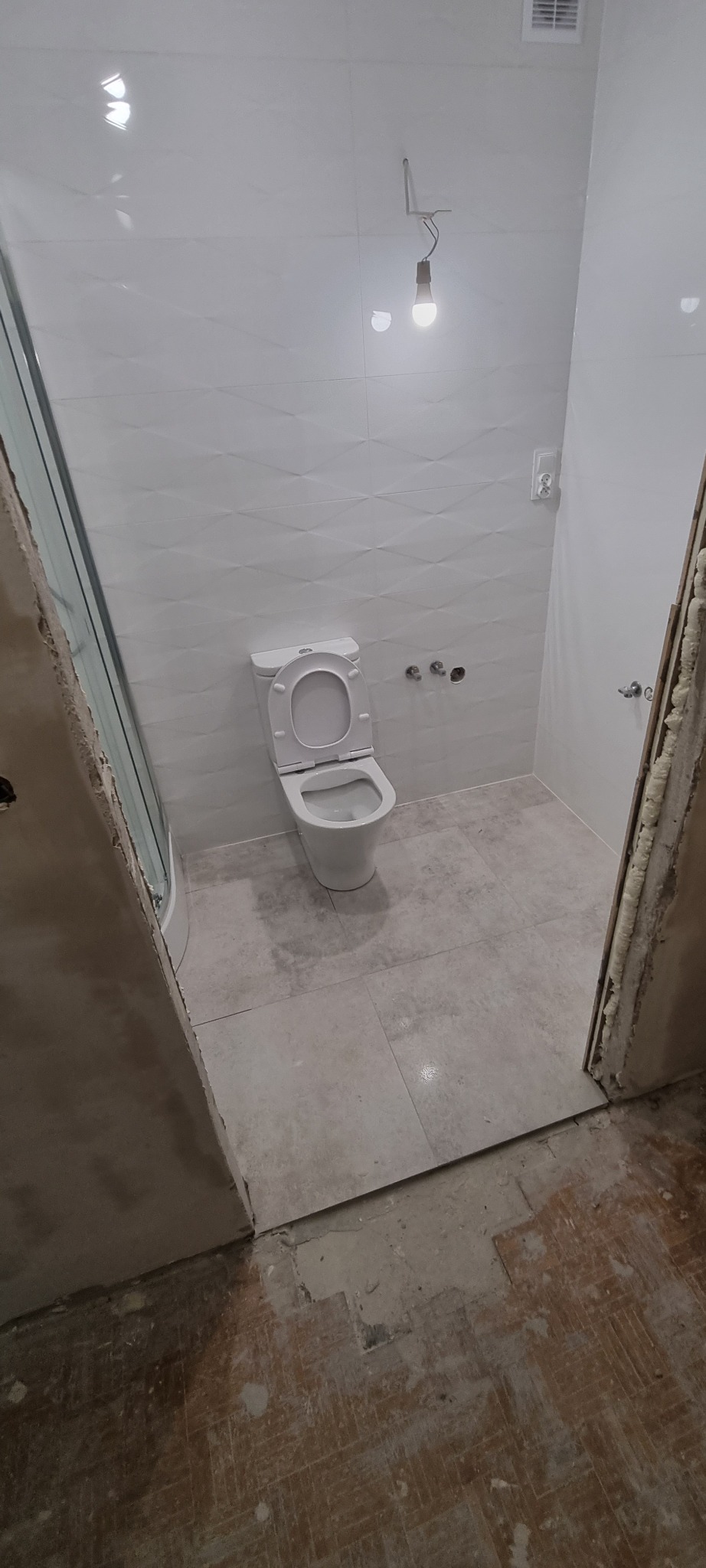 Biała, częściowo wykończona łazienka z toaletą, widoczne płytki na ścianach i podłodze, surowy stan podłogi przed wejściem do łazienki, tymczasowa żarówka na kablu.