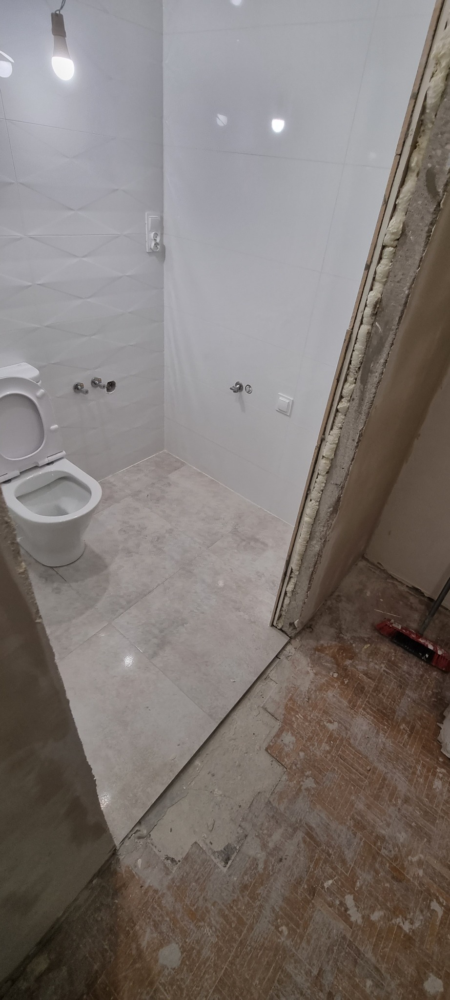 Łazienka w trakcie remontu: położone płytki podłogowe i ścienne, widoczny nowy sedes, przygotowane otwory na armaturę, pianka montażowa wokół ościeżnicy, stara drewniana podłoga poza wyremontowaną...