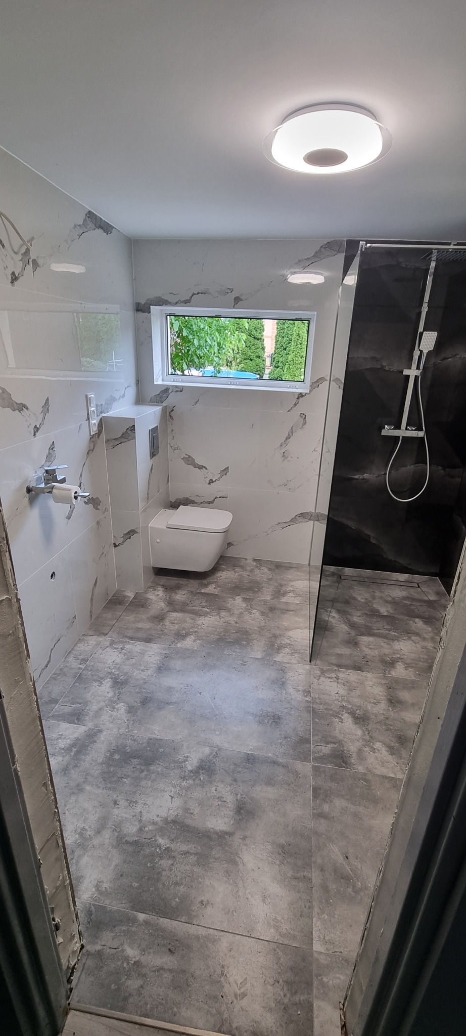 Nowoczesna łazienka z marmurowymi płytkami na ścianach, szarą podłogą imitującą beton, podwieszaną toaletą i kabiną prysznicową typu walk-in z czarnym panelem.