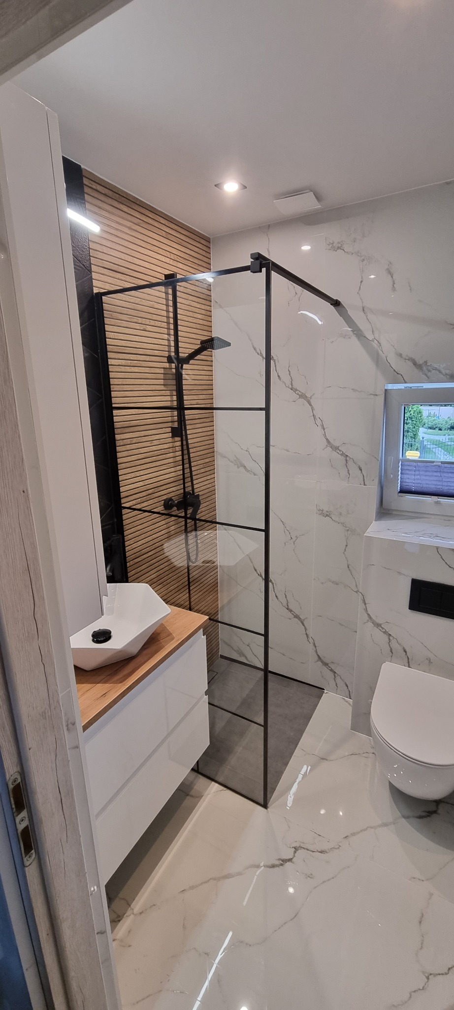 Nowoczesna łazienka z kabiną prysznicową typu walk-in z czarnymi profilami, umywalką nablatową o nietypowym kształcie i drewnianym blatem, oraz toaletą. Na ścianach płytki imitujące marmur...