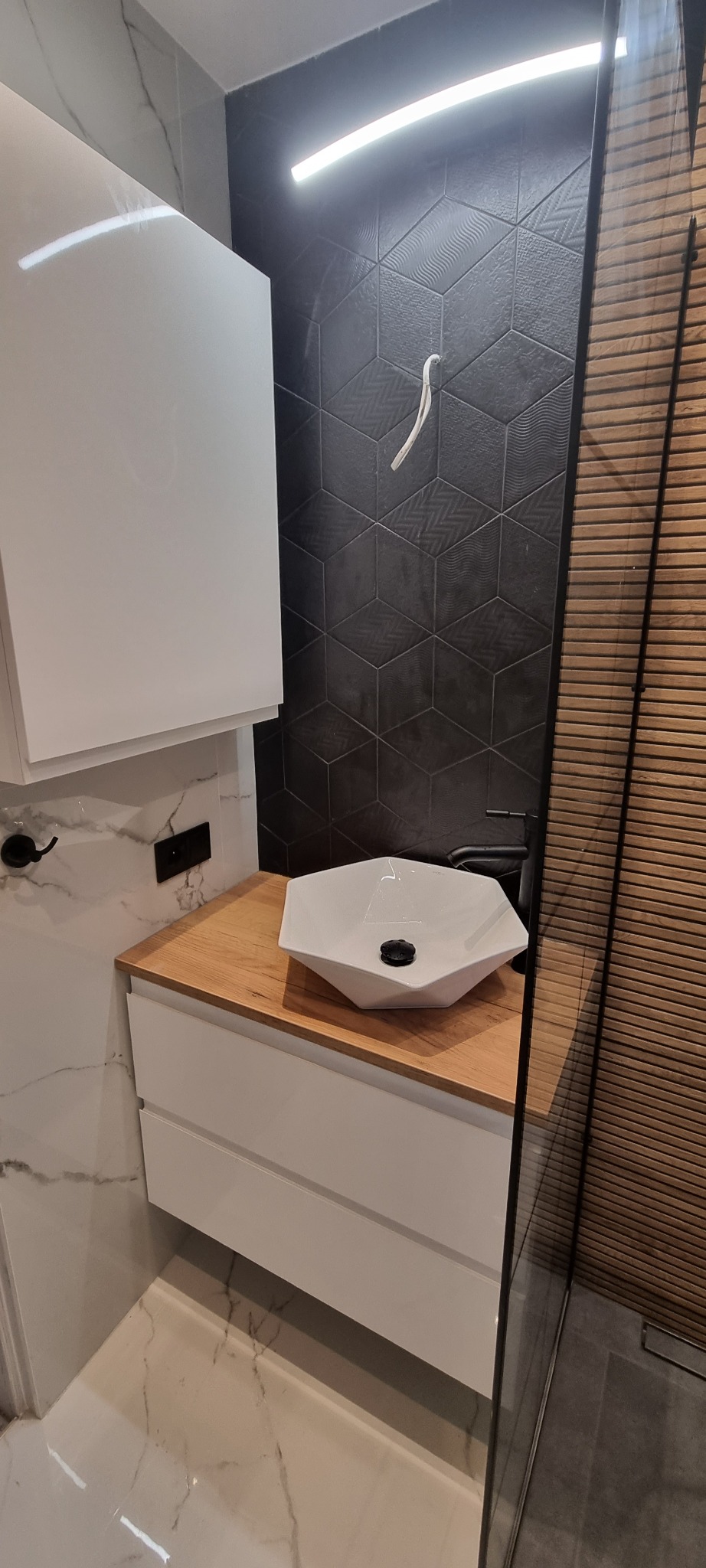 Nowoczesna łazienka: biała szafka z umywalką w kształcie wielokąta na drewnianym blacie, czarna armatura, heksagonalne płytki na ścianie i drewniane lamele prysznicowe.