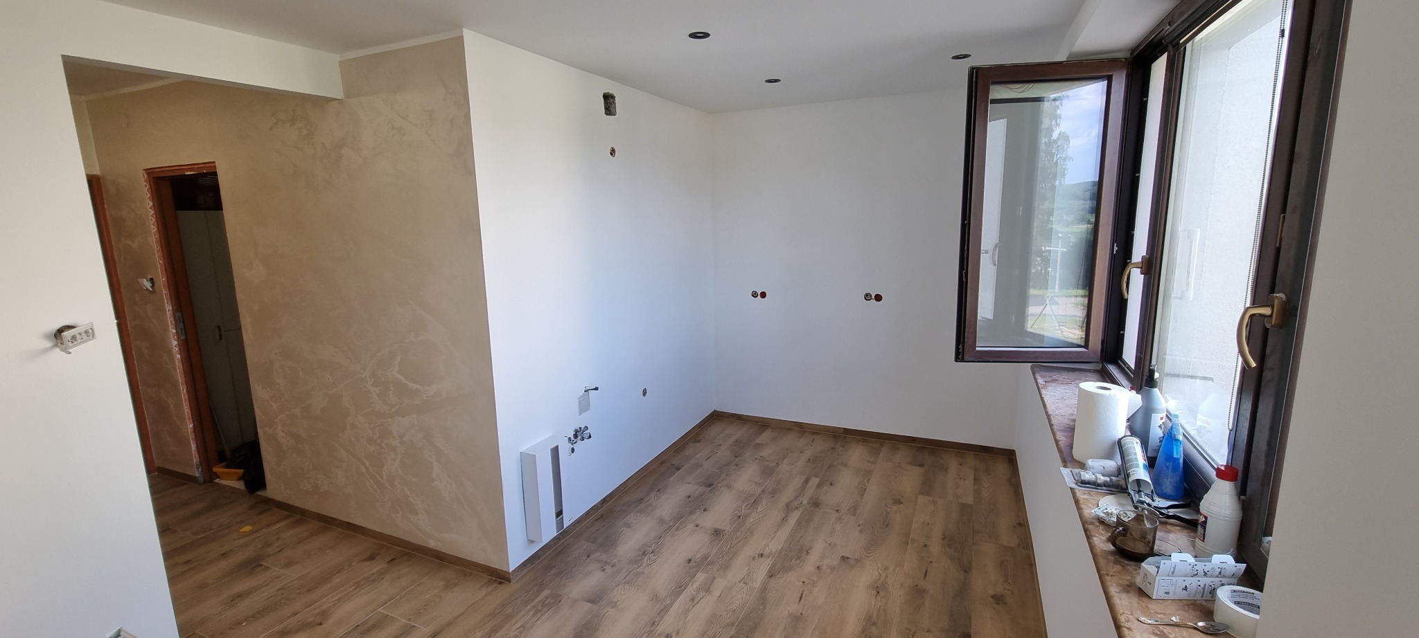 Wnętrze pokoju w trakcie prac wykończeniowych: pomalowane ściany na biało, podłoga wyłożona panelami imitującymi drewno, częściowo wykończona ściana z efektem dekoracyjnym, otwarte okno...