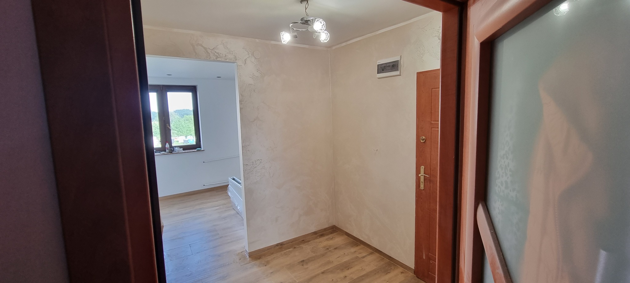 Wnętrze mieszkania po remoncie: widok na odnowione ściany z efektem dekoracyjnym, drewnopodobne panele na podłodze, nowe drzwi wewnętrzne i widok na pokój z oknem.