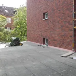 Prace przygotowawcze na dachu budynku z czerwoną ceglaną elewacją, widoczne rusztowanie, wiadro z farbą, narzędzia oraz materiały budowlane pod plandeką.