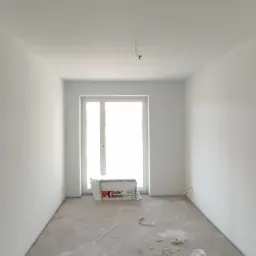 Wnętrze pokoju po malowaniu ścian na biało, widoczne opakowanie materiałów izolacyjnych przy oknie, surowa podłoga, wystające przewody z sufitu.