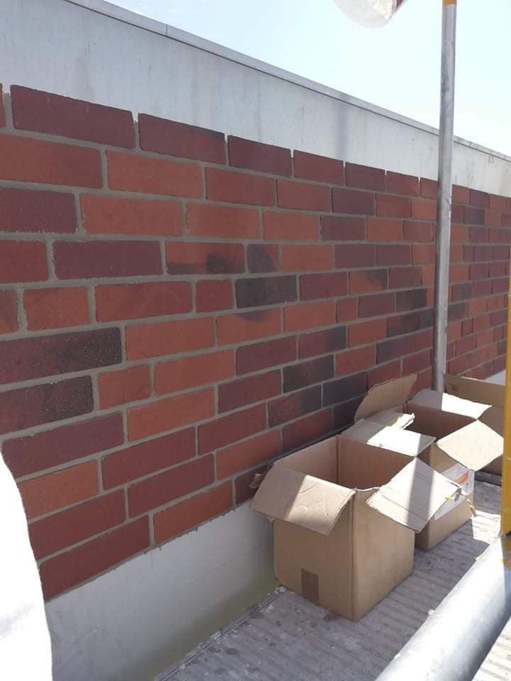 Fragment elewacji z czerwonej cegły klinkierowej w trakcie montażu, widoczne niedokończone spoiny i kartony na rusztowaniu.
