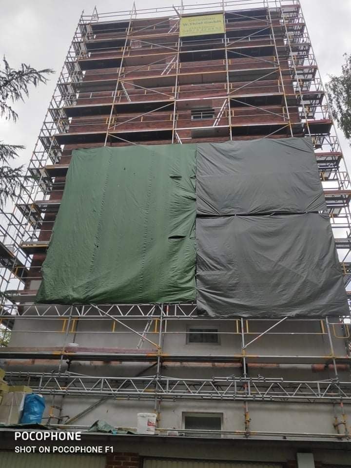 Wielopiętrowy budynek w trakcie prac elewacyjnych, osłonięty częściowo zieloną i szarą plandeką, z widocznym rusztowaniem i żółtą tablicą informacyjną na szczycie.