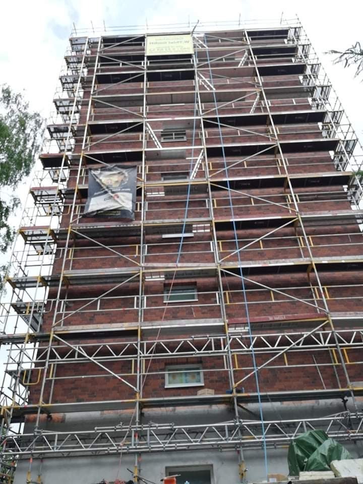 Wielopiętrowy budynek z czerwonej cegły w trakcie prac elewacyjnych, otoczony rusztowaniem, widoczny baner reklamowy na rusztowaniu.