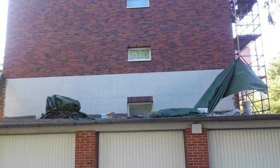 Częściowo otynkowana ściana budynku z czerwonej cegły, widoczne rusztowanie, zabezpieczenia folią i garaże w dolnej części.