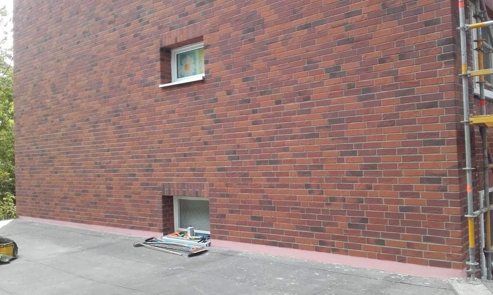 Murowana elewacja budynku z cegły klinkierowej w odcieniach czerwieni i brązu, z widocznymi rusztowaniami i narzędziami na dachu, wykonana z dbałością o detale.