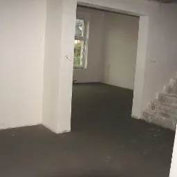 Świeżo wylana, gładka posadzka w surowym wnętrzu domu z widocznymi schodami w trakcie wykończenia.