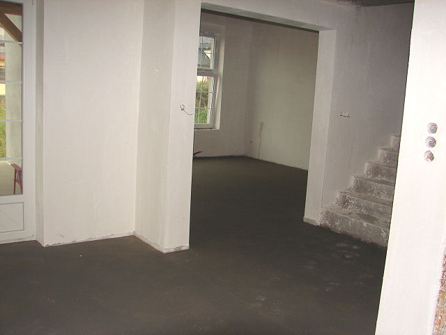 Świeżo wylana, gładka posadzka w surowym wnętrzu domu z widocznymi schodami w trakcie wykończenia.