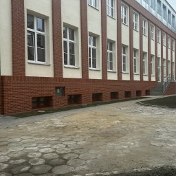 MAXIBUDD - Odnowiona elewacja budynku z ceglaną podstawą i białymi pasami, widoczne okna z białymi ramami i metalowe elementy dachu. Uwagę przykuwa wzór chodnika.