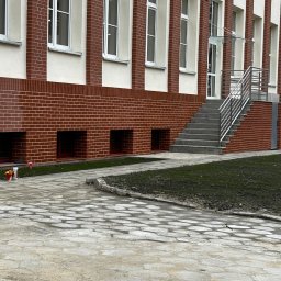 MAXIBUDD - Budynek z ceglaną elewacją, białymi oknami i schodami z metalową barierką. Widoczne detale wykończenia i otoczenie budynku.