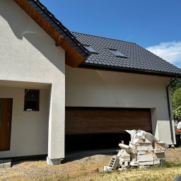 MAXIBUDD - Nowoczesny dom z jasną elewacją i drewnianą bramą garażową. Na pierwszym planie stos białych bloczków, obok stoi mężczyzna. Błękitne niebo w tle.