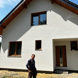 MAXIBUDD - Biały dom jednorodzinny z ciemnym dachem i drewnianymi oknami. Na pierwszym planie mężczyzna w ubraniu roboczym. Widoczne świeżo położone tynki.