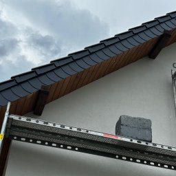 MAXIBUDD - Fragment elewacji domu z szarą dachówką, widoczny ze rusztowania. Ściana otynkowana na biało, pochmurne niebo w tle. Detal prac elewacyjnych.