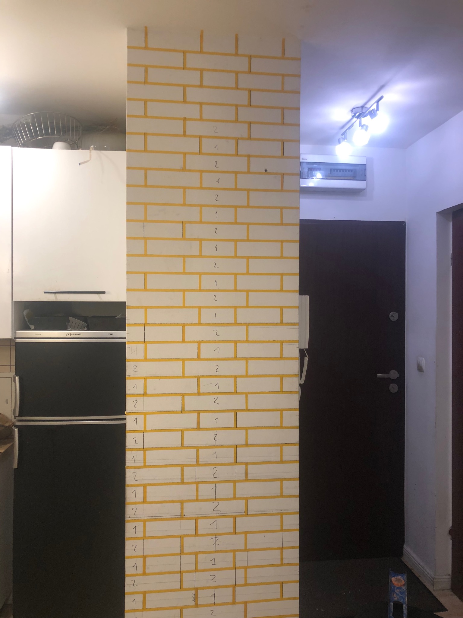 Wnętrze z fragmentem ściany imitującej cegłę, z żółtymi fugami i numeracją. Obok widoczna zabudowa kuchenna i drzwi wejściowe.