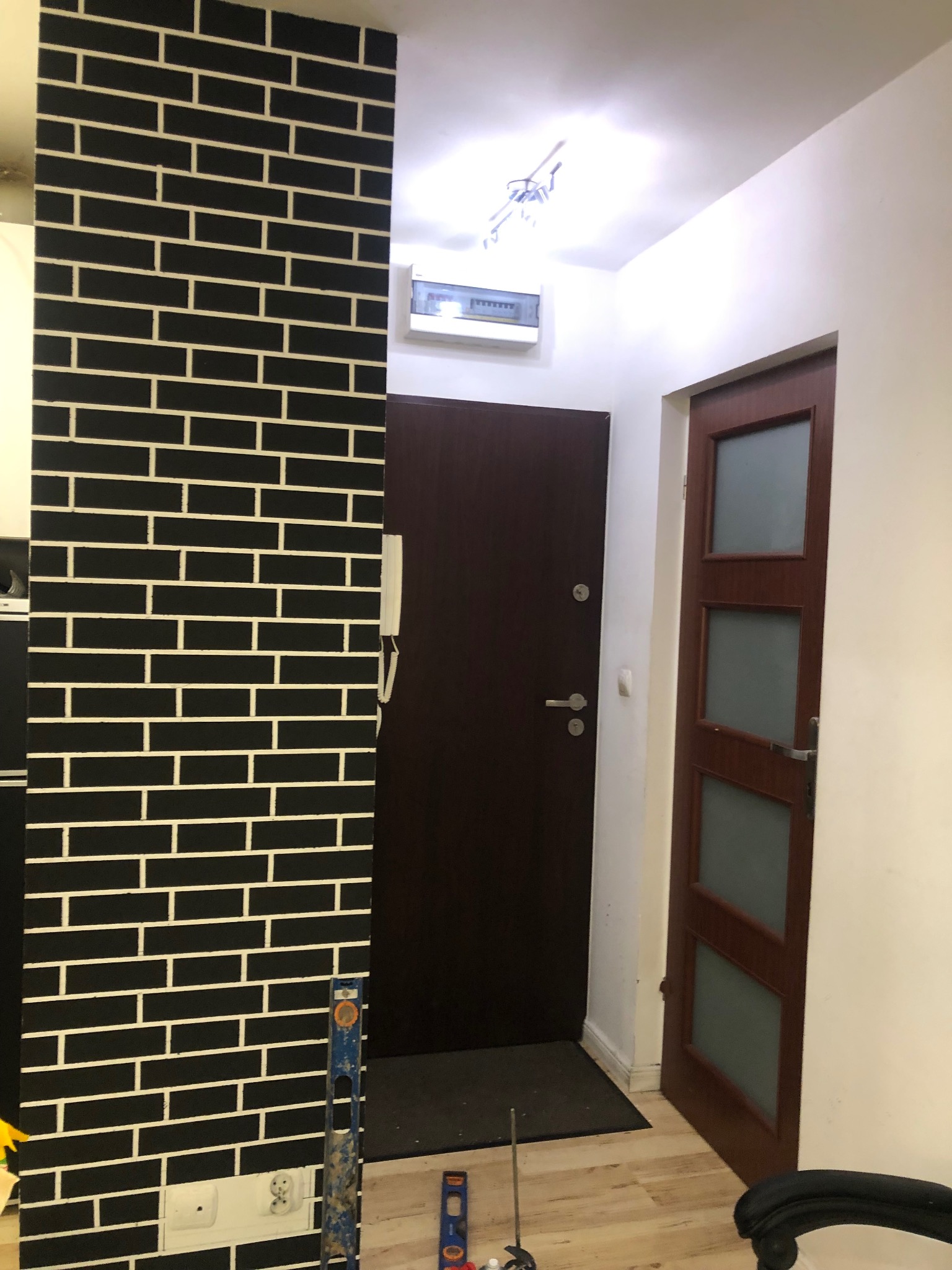 Ściana w korytarzu wykończona czarną cegłą z białymi fugami, obok drzwi wejściowe i wewnętrzne. Widoczne narzędzia pomiarowe.