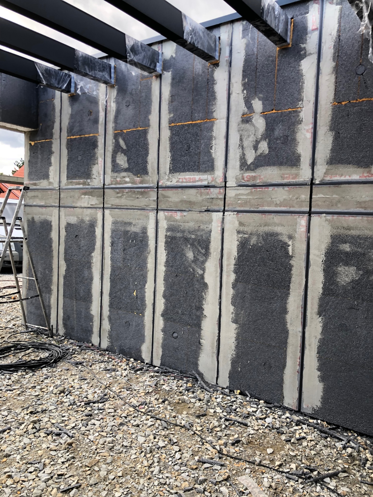 Szara ściana budynku w trakcie montażu paneli izolacyjnych z widocznymi spoinami i siatką wzmacniającą, na żwirowym podłożu, z drabiną i elementami konstrukcji dachu.
