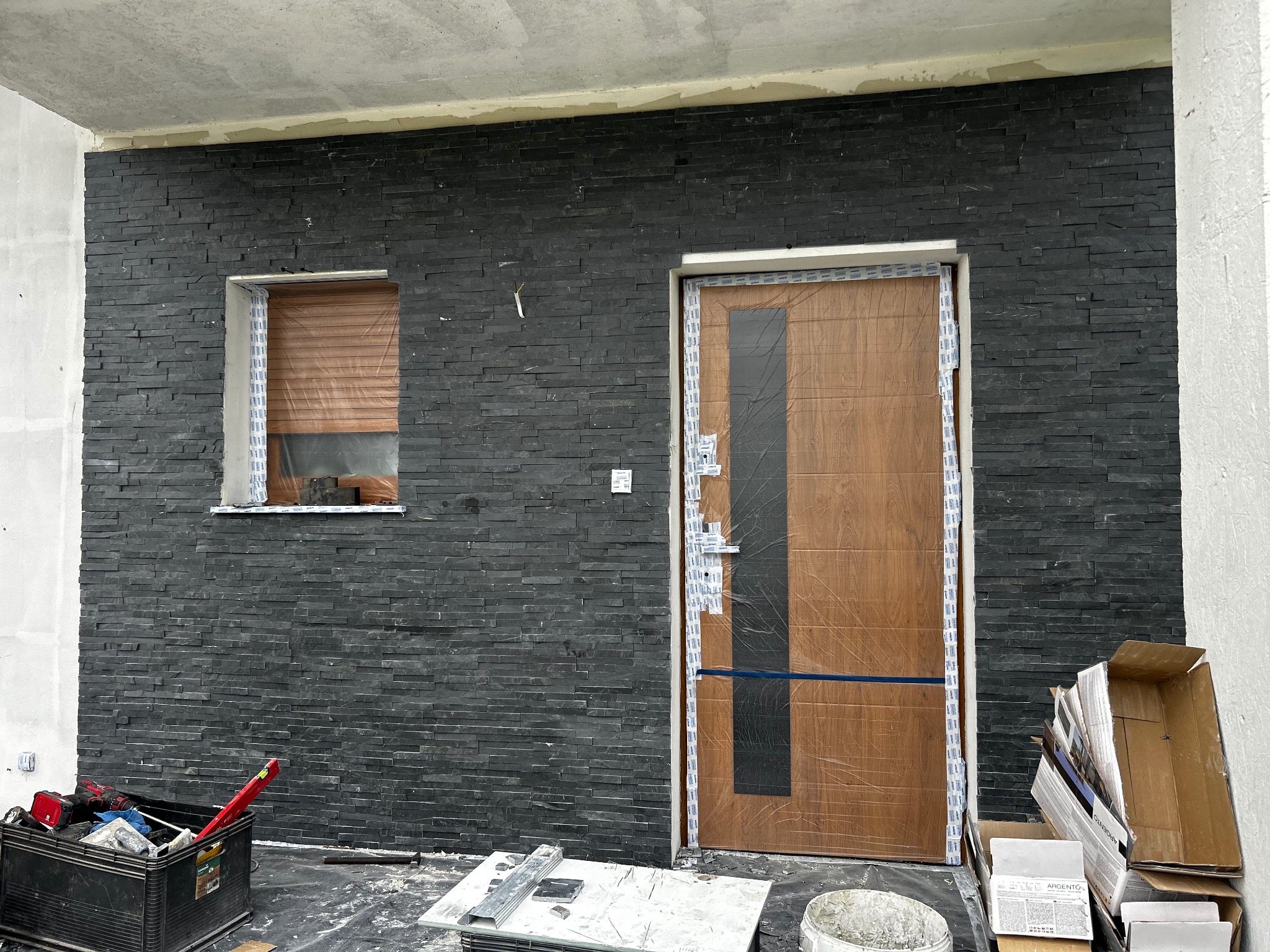 Elewacja z czarnego kamienia dekoracyjnego, okno z brązową roletą, drewniane drzwi zabezpieczone folią. Widoczne narzędzia i materiały budowlane.
