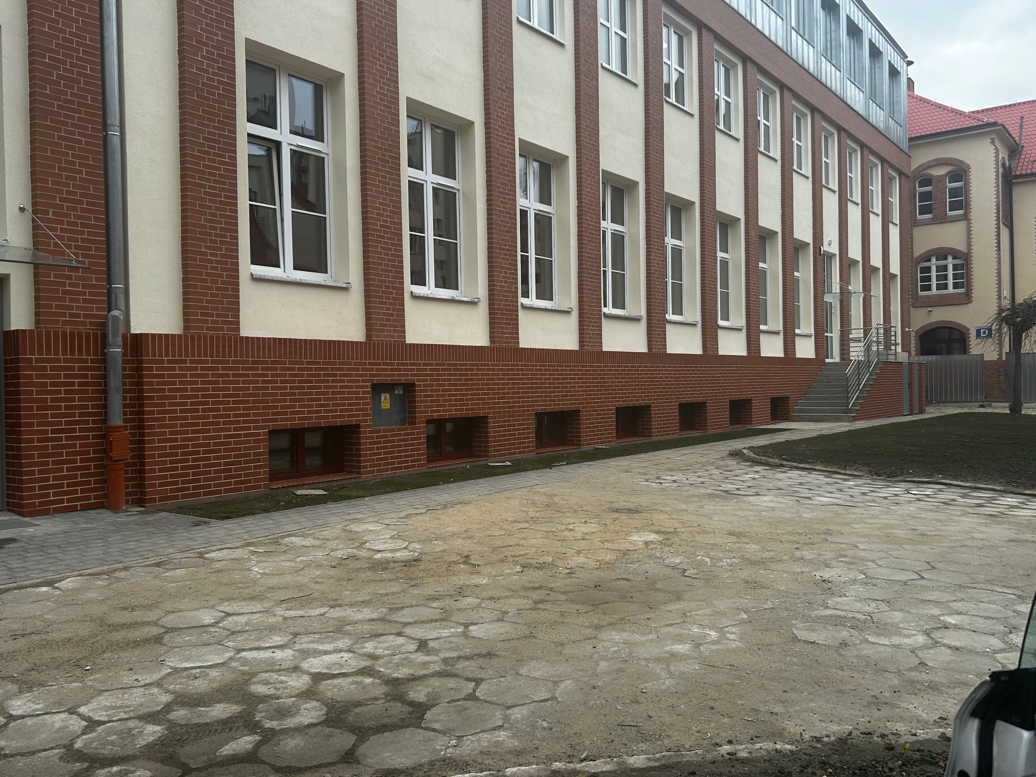 Odnowiona elewacja budynku z ceglaną podstawą i białymi pasami, widoczne okna z białymi ramami i metalowe elementy dachu. Uwagę przykuwa wzór chodnika.