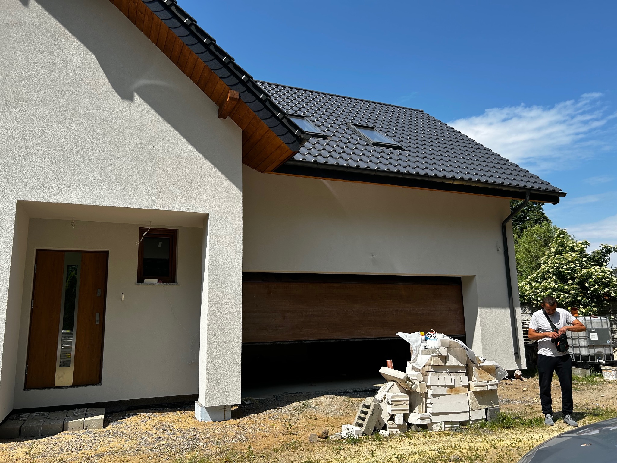 Nowoczesny dom z jasną elewacją i drewnianą bramą garażową. Na pierwszym planie stos białych bloczków, obok stoi mężczyzna. Błękitne niebo w tle.