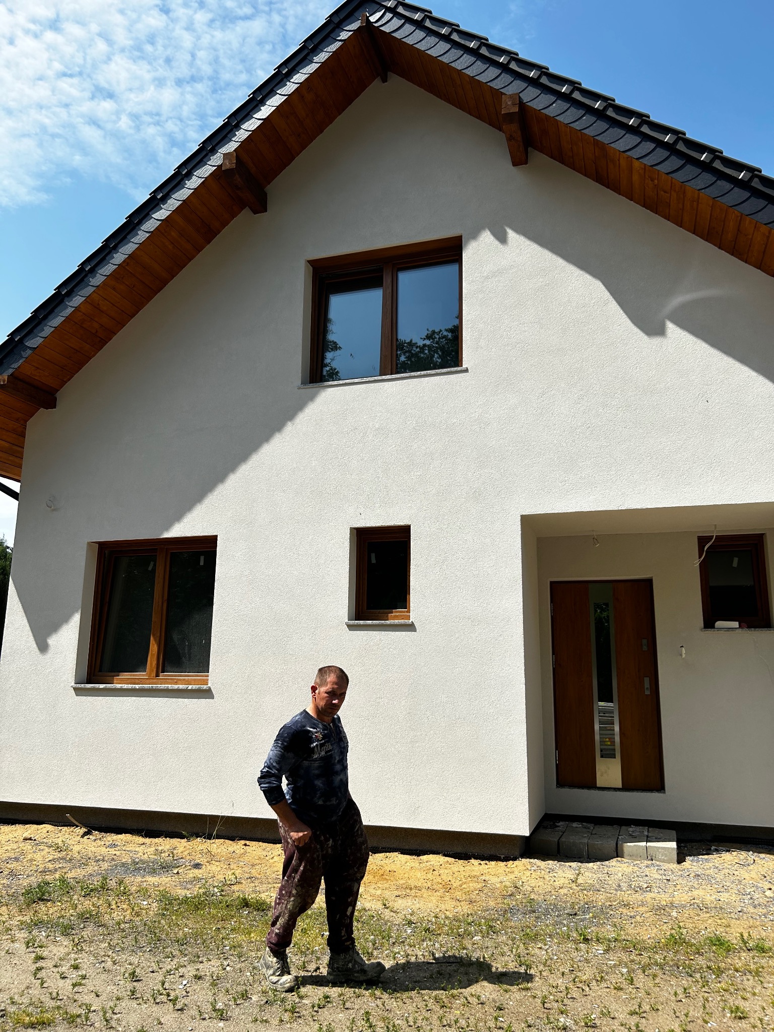 Biały dom jednorodzinny z ciemnym dachem i drewnianymi oknami. Na pierwszym planie mężczyzna w ubraniu roboczym. Widoczne świeżo położone tynki.