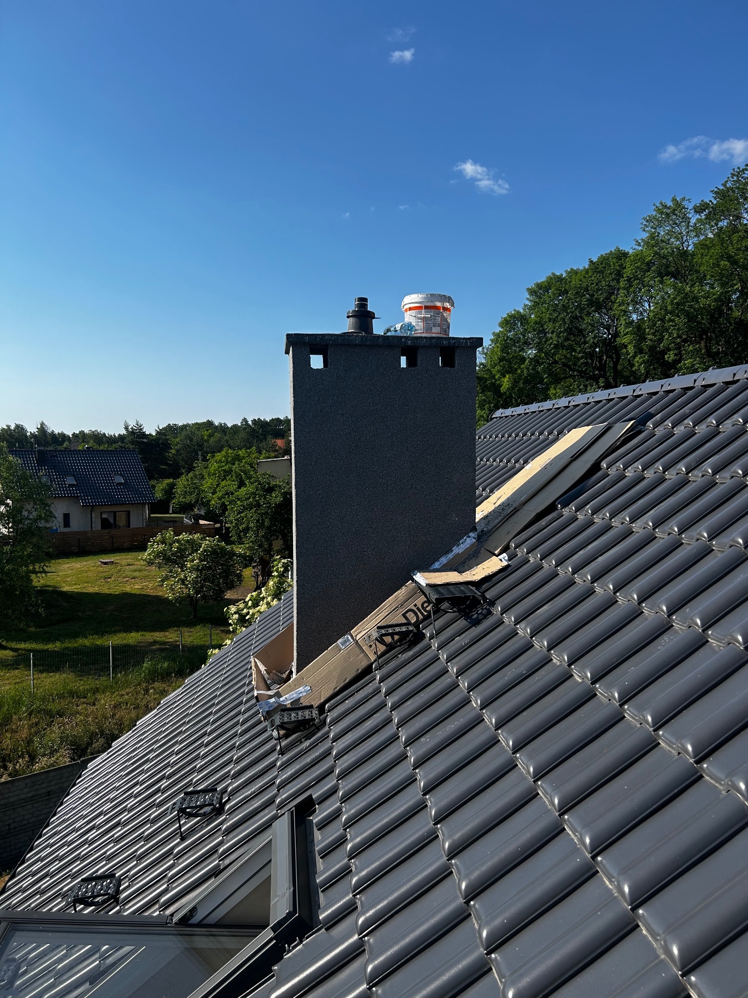 Szare dachówki ceramiczne na dachu domu we Wrocławiu, widok z góry na komin i fragment dachu w trakcie prac dekarskich, błękitne niebo w tle.