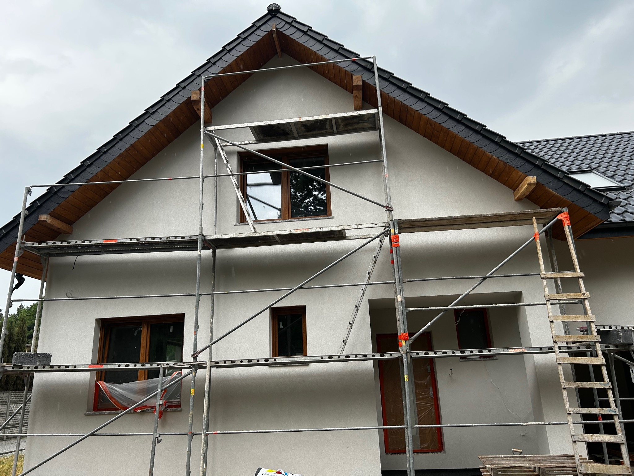 Dom jednorodzinny w trakcie prac elewacyjnych, widoczne rusztowania i zabezpieczone okna. Świeżo otynkowana, jasna elewacja kontrastuje z ciemną dachówką.