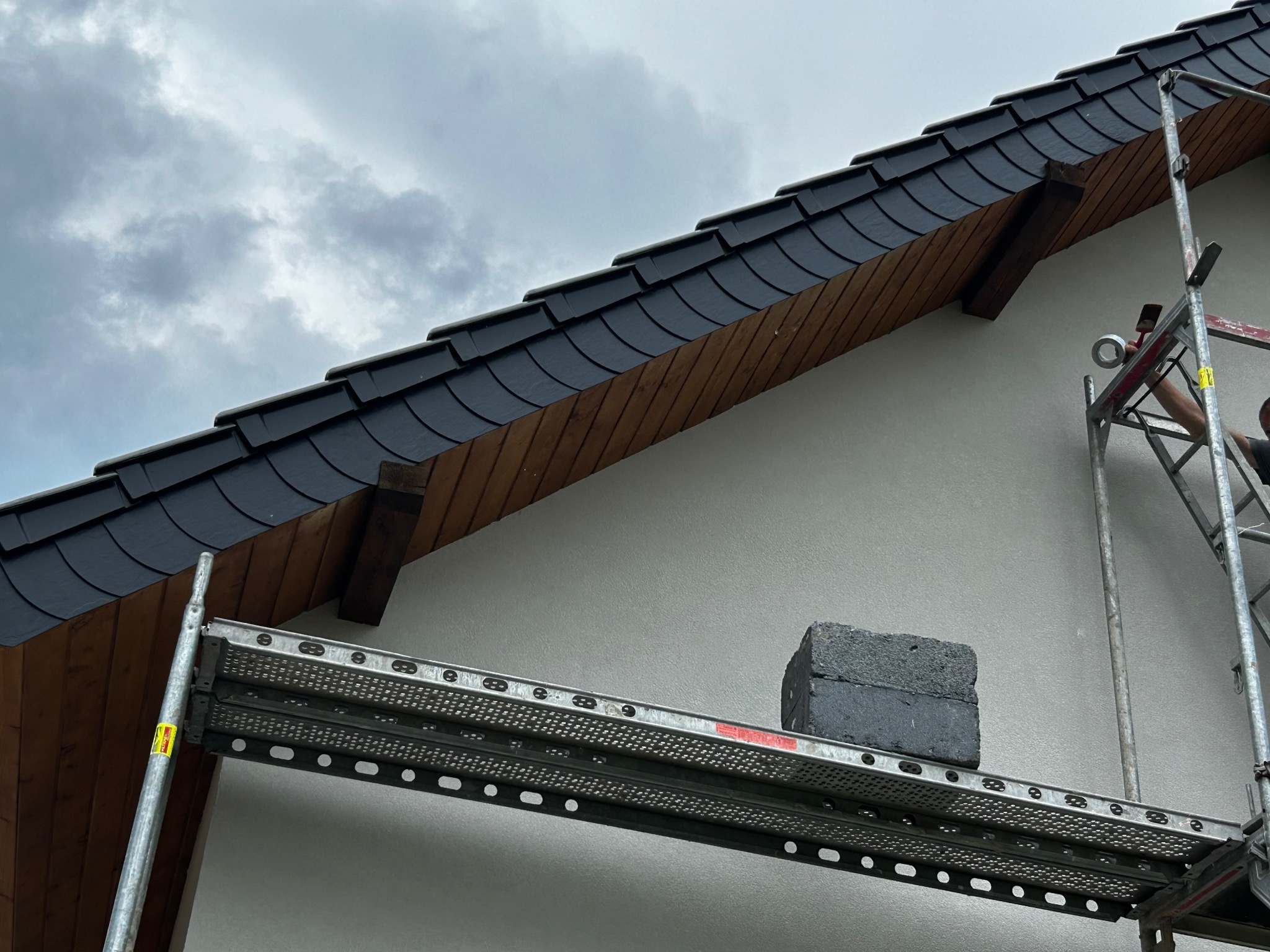 Fragment elewacji domu z szarą dachówką, widoczny ze rusztowania. Ściana otynkowana na biało, pochmurne niebo w tle. Detal prac elewacyjnych.