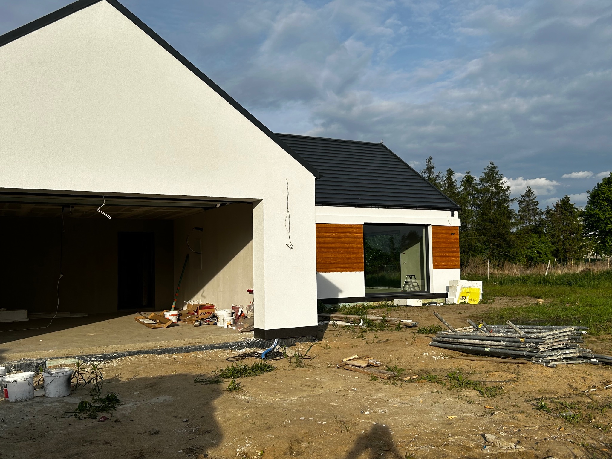 Nowoczesny dom jednorodzinny z białą elewacją i drewnianymi akcentami, w trakcie wykańczania. Widoczne rusztowania i materiały budowlane. Dom z garażem w surowym stanie.