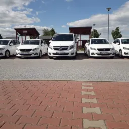 Flota białych samochodów osobowych i busów zaparkowana rzędem na parkingu z kostki brukowej, widoczne tablice rejestracyjne z Warszawy.