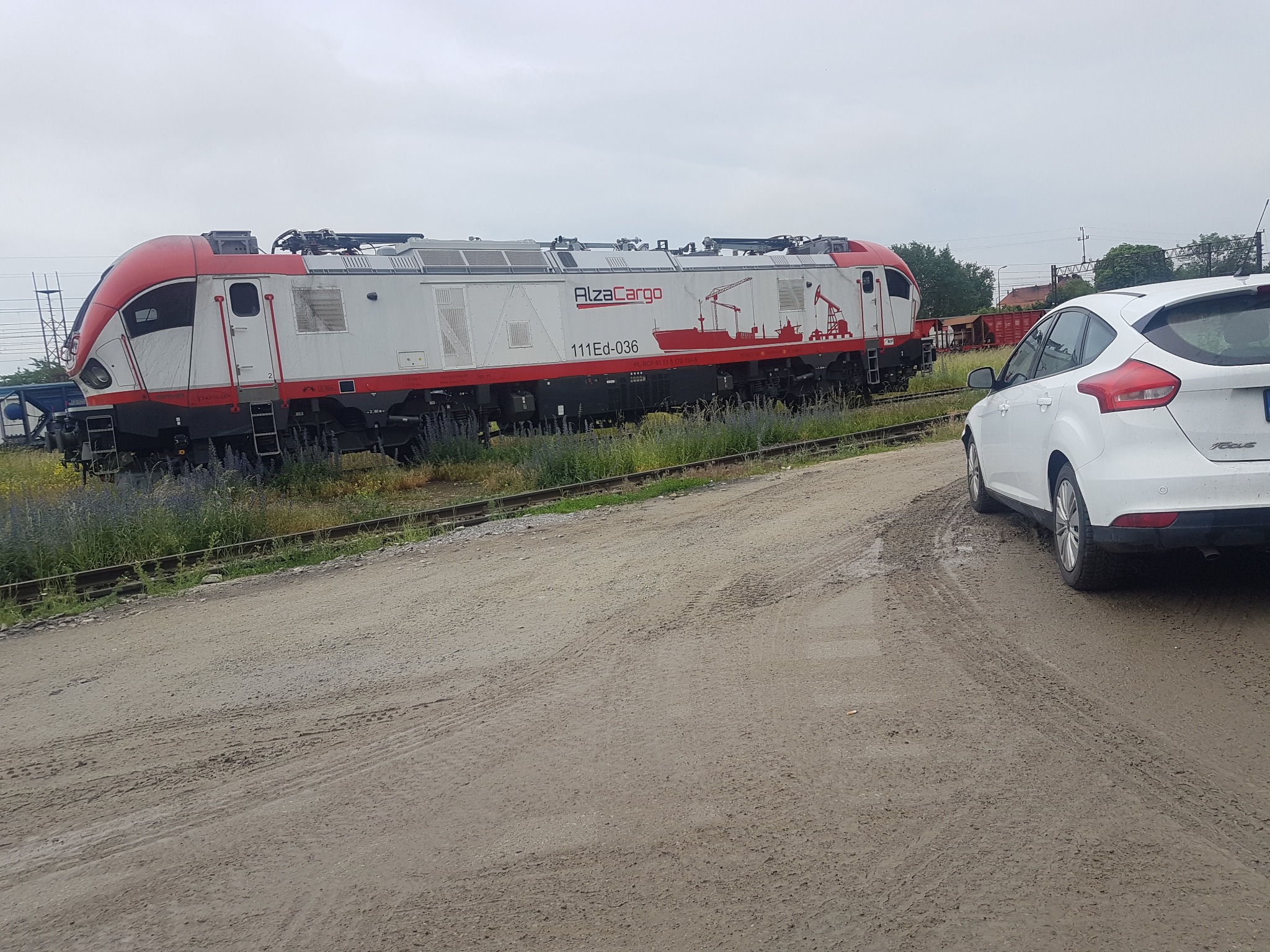 Elektryczna lokomotywa Alza Cargo 111Ed-036 z wagonami w tle, widziana obok zaparkowanego białego samochodu osobowego na piaszczystej drodze.